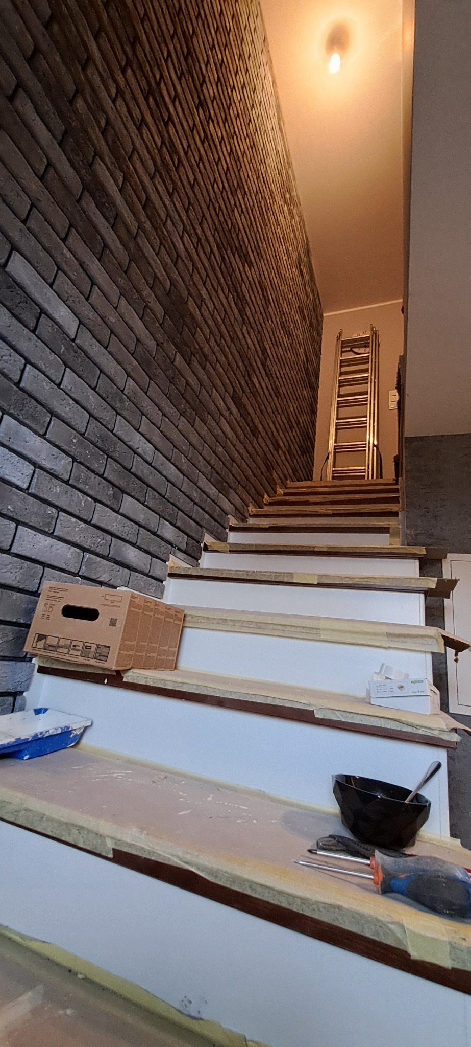 Schody w trakcie wykończenia, zabezpieczone taśmą malarską, z widoczną ścianą wyłożoną cegłą dekoracyjną, narzędzia i drabina w tle.