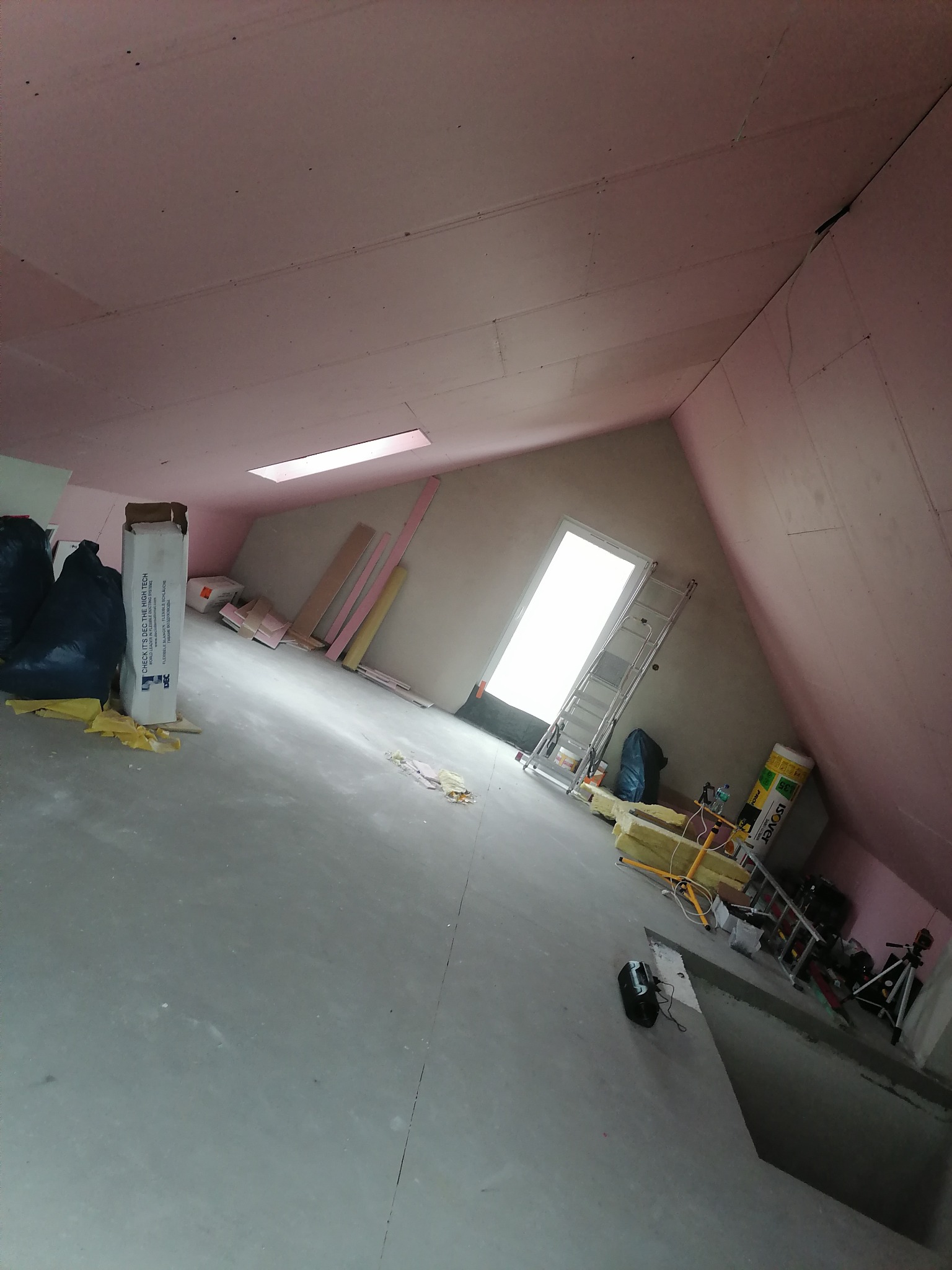 Poddasze w trakcie prac ociepleniowych: widoczne płyty izolacyjne na ścianach i suficie, materiały budowlane, drabina i narzędzia pomiarowe. Na podłodze rozłożona żółta wełna mineralna. Ujęcie...