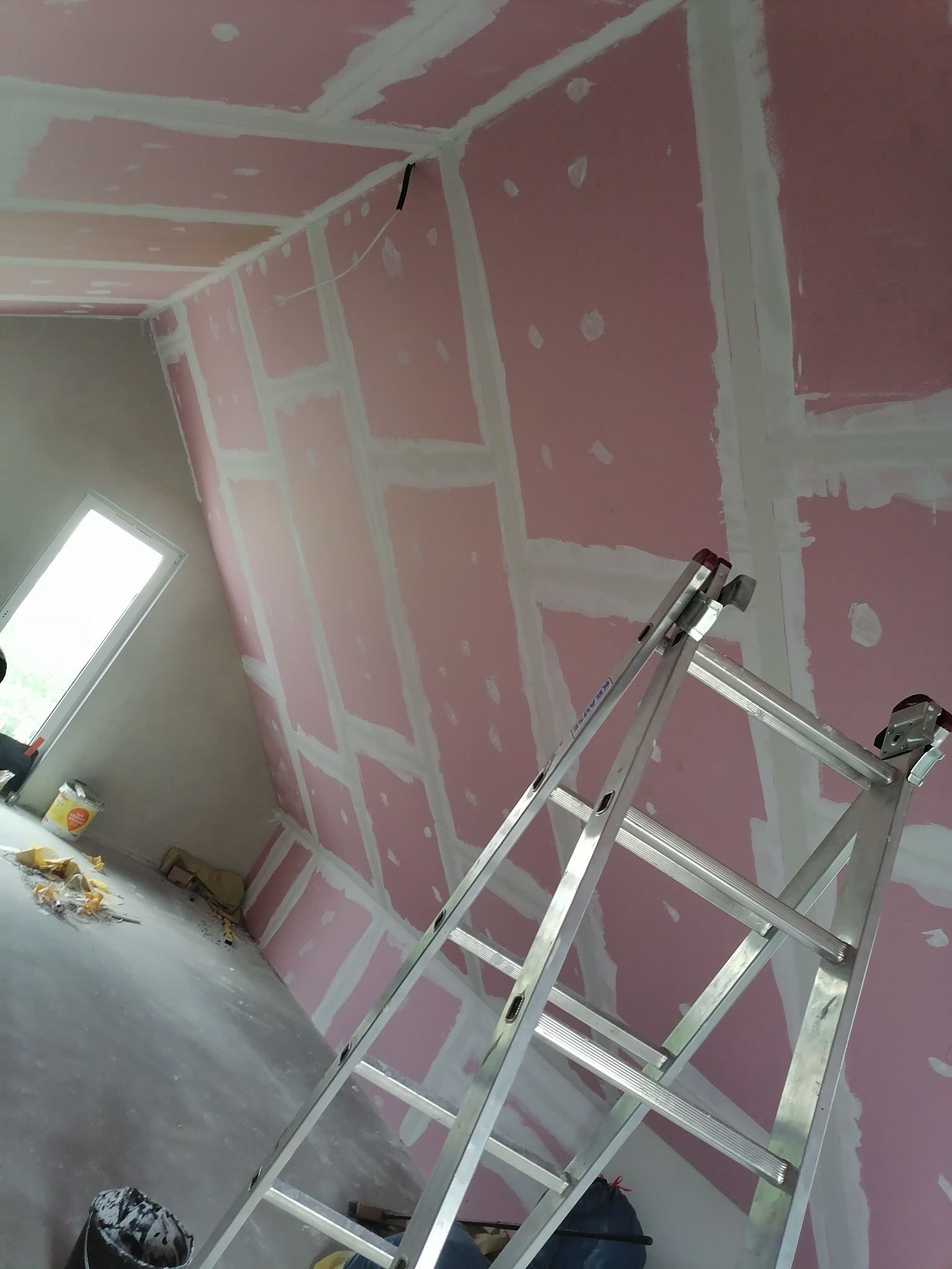 Poddasze w trakcie wykańczania: płyty gipsowo-kartonowe z widocznymi łączeniami pokrytymi gładzią szpachlową, aluminiowa drabina rozstawiona w pomieszczeniu, okno dachowe w tle.