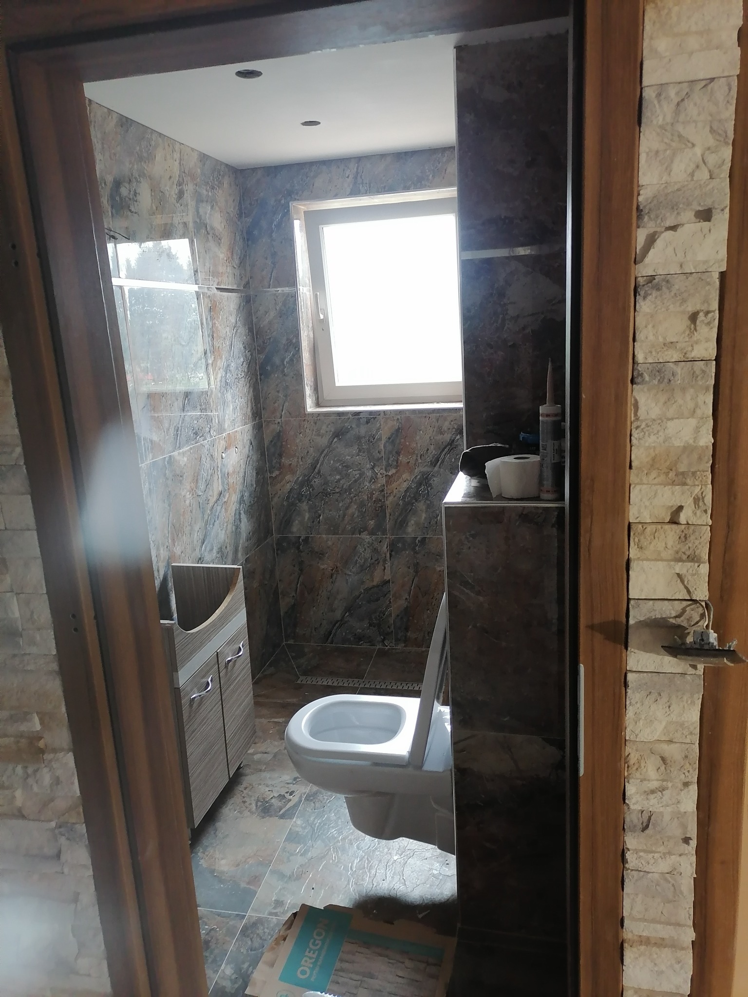 Wykończona łazienka z brązowo-szarymi płytkami imitującymi marmur na ścianach i podłodze, widoczna umywalka z szafką, toaleta i okno. Dekoracyjne wykończenie ościeżnicy z kamienia elewacyjnego.