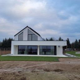 BP Construction BARTŁOMIEJ POLAK - Nowoczesny, biały dom z dużymi oknami i czarnym dachem dwuspadowym, otoczony świeżo założonym trawnikiem i drzewami iglastymi, sfotografowany w pochmurny dzień.