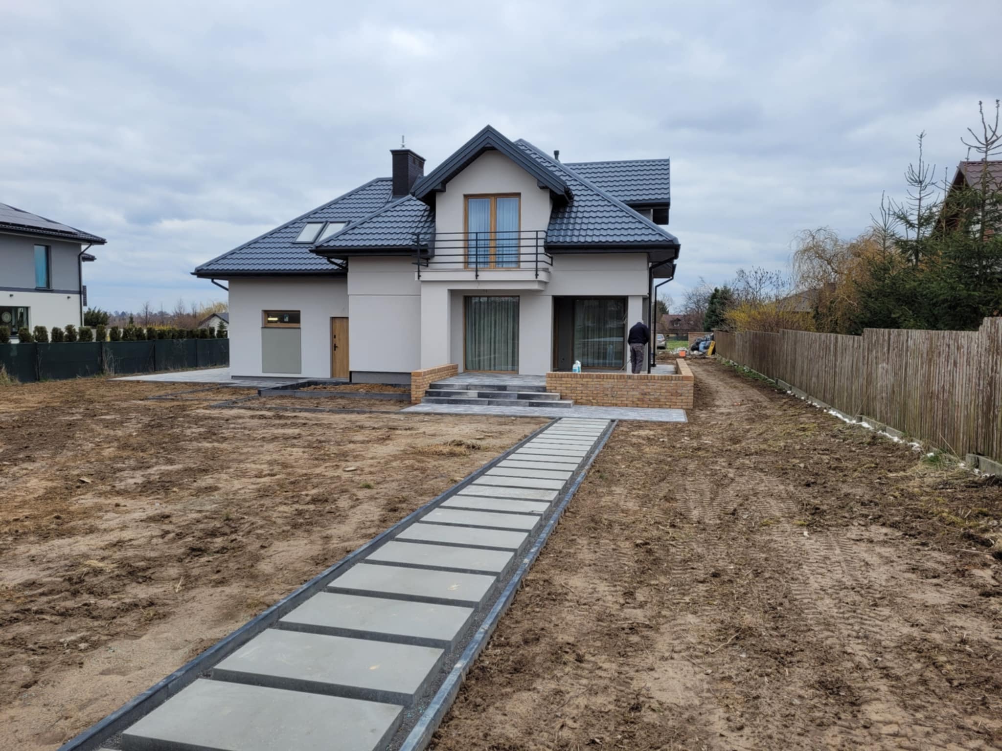 Nowoczesny dom jednorodzinny z szarą dachówką i jasną elewacją. Ułożony chodnik z płyt prowadzi do wejścia. Widoczne prace wykończeniowe wokół budynku.
