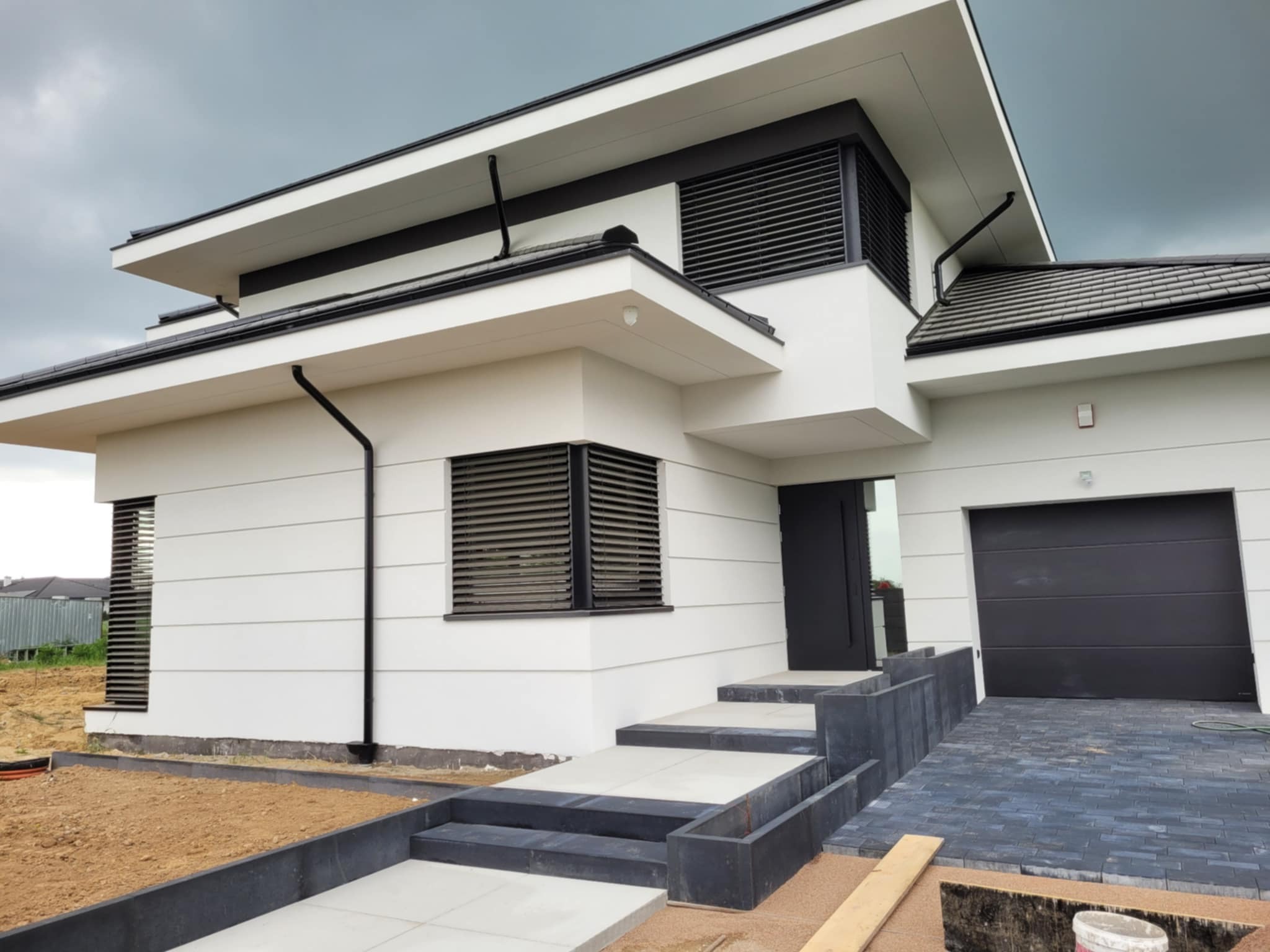 Nowoczesny dom jednorodzinny z płaskim dachem, białymi ścianami i grafitowymi detalami, w tym żaluzjami fasadowymi i bramą garażową. Uwagę przykuwa minimalistyczny design.