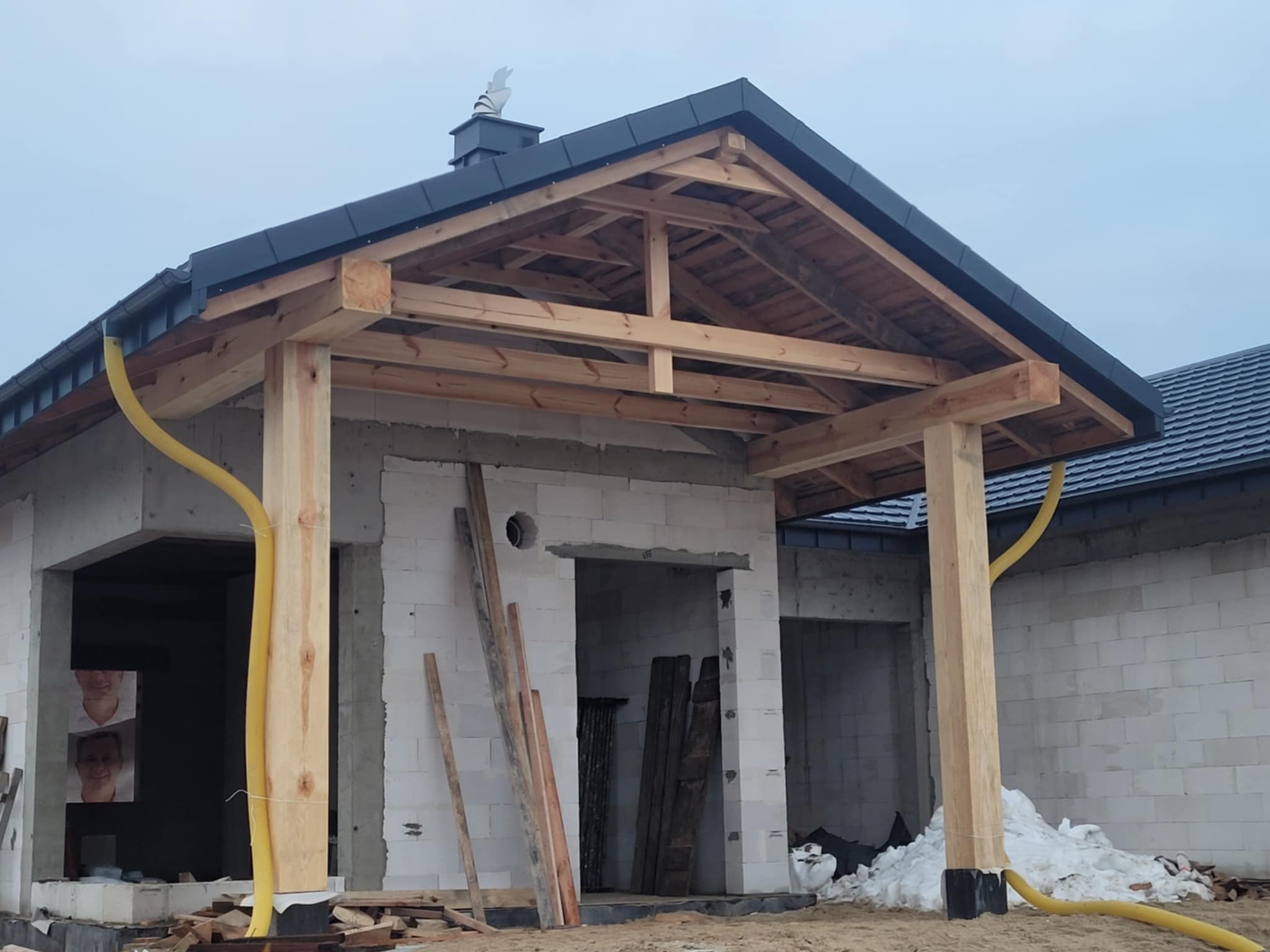 Surowy stan zadaszenia tarasu z drewnianą konstrukcją i częściowo położonym dachem z dachówki, widoczne materiały budowlane i przewody wentylacyjne.