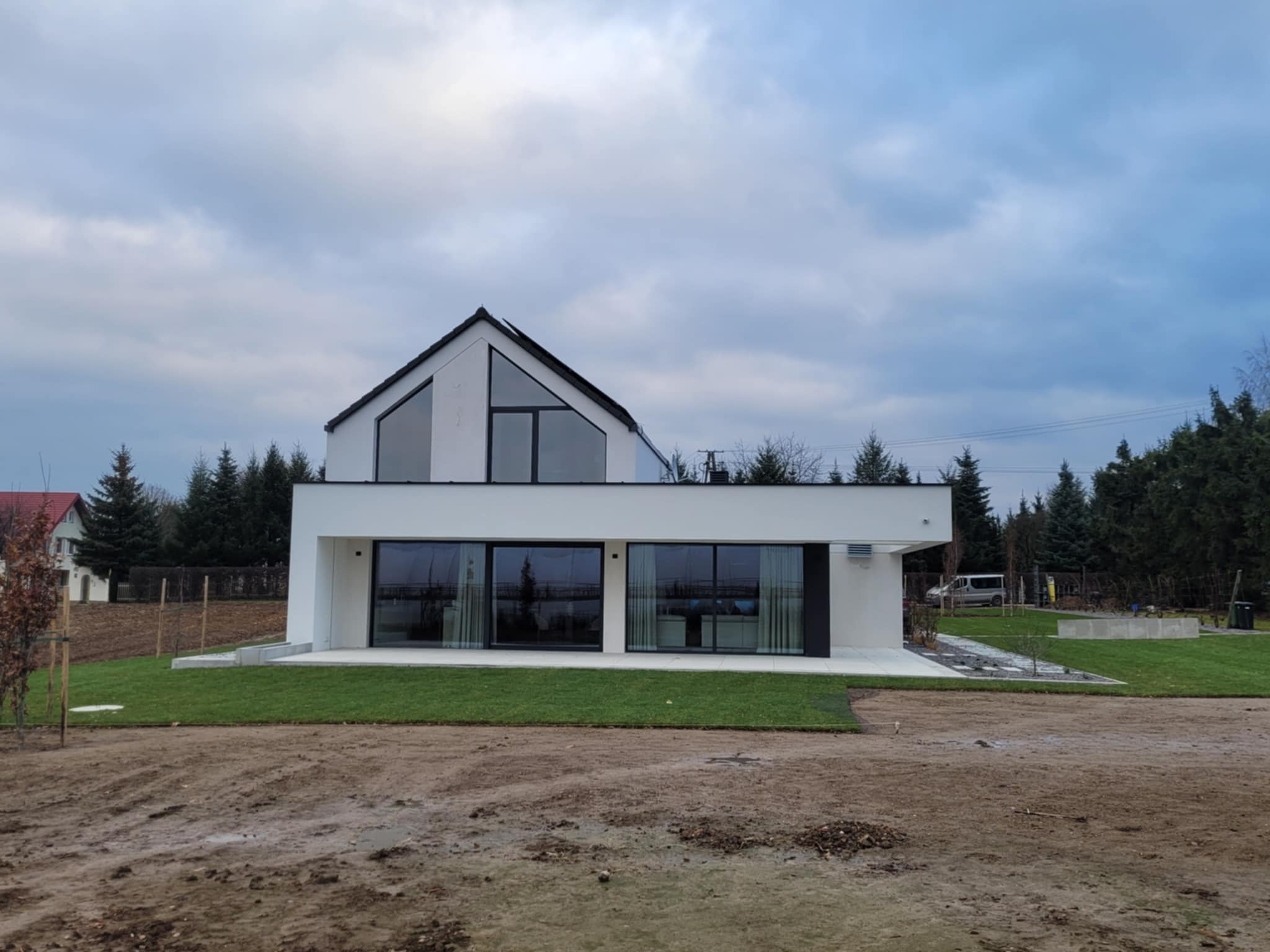 Nowoczesny, biały dom z dużymi oknami i czarnym dachem dwuspadowym, otoczony świeżo założonym trawnikiem i drzewami iglastymi, sfotografowany w pochmurny dzień.