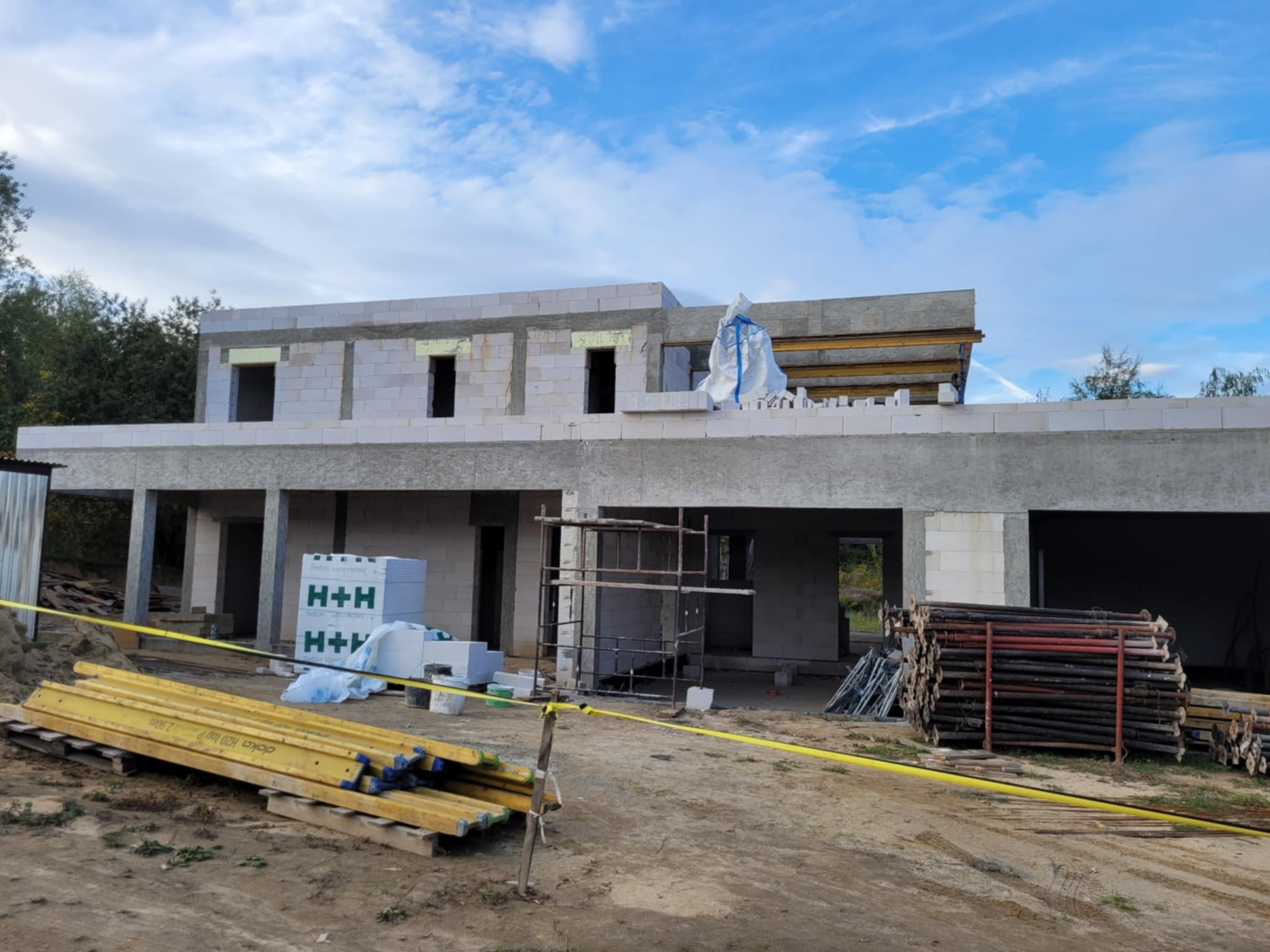 Budowa domu jednorodzinnego w stanie surowym otwartym, widoczne bloczki H+H, szalunki, rusztowania i materiały budowlane. Ujęcie w słoneczny dzień.