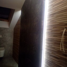 EKOMARBUD - Nowoczesna łazienka w trakcie wykańczania: ciemna ściana z efektem betonu kontrastuje z drewnianymi panelami na ścianie, oświetlonymi listwą LED. Widoczny fragment toalety i okno dachowe.