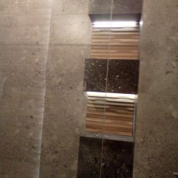 EKOMARBUD - Fragment ściany łazienki z szarą, nakrapianą płytką oraz pionowy pas z ciemnej płytki z jasnymi plamkami, podświetlony od góry, z widocznymi drewnianymi żaluzjami w tle.