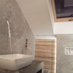 EKOMARBUD - Łazienka w trakcie remontu: widoczna biała umywalka na ciemnym blacie, szara ściana z instalacją elektryczną, drewniane lamele w tle i okno dachowe.