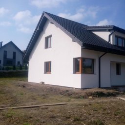 EKOMARBUD - Nowo ocieplony dom jednorodzinny z białymi ścianami i ciemną dachówką, widoczne brązowe ramy okienne, teren wokół domu jeszcze niezagospodarowany.