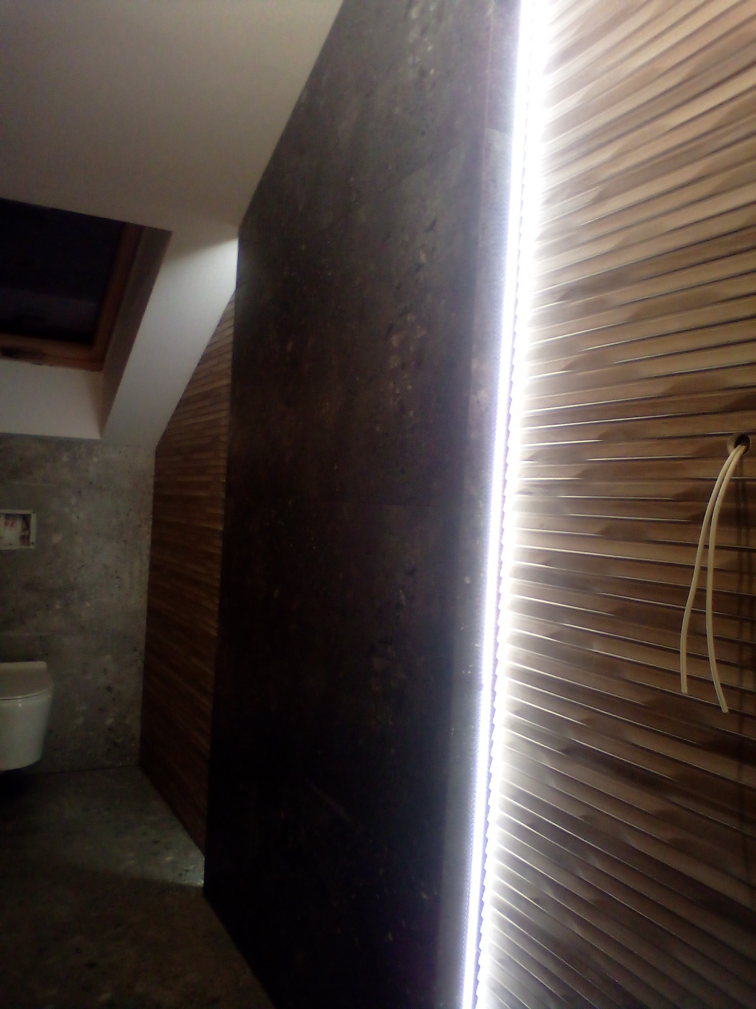 Nowoczesna łazienka w trakcie wykańczania: ciemna ściana z efektem betonu kontrastuje z drewnianymi panelami na ścianie, oświetlonymi listwą LED. Widoczny fragment toalety i okno dachowe.