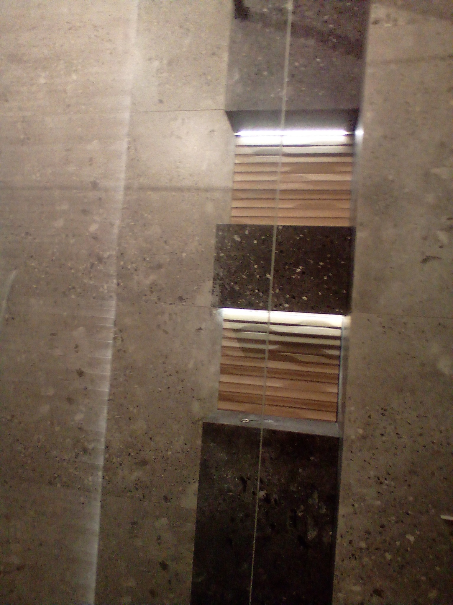 Fragment ściany łazienki z szarą, nakrapianą płytką oraz pionowy pas z ciemnej płytki z jasnymi plamkami, podświetlony od góry, z widocznymi drewnianymi żaluzjami w tle.
