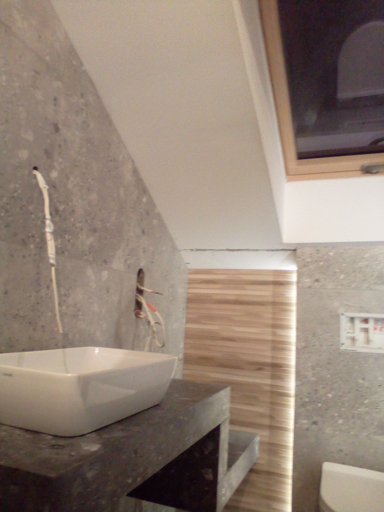 Łazienka w trakcie remontu: widoczna biała umywalka na ciemnym blacie, szara ściana z instalacją elektryczną, drewniane lamele w tle i okno dachowe.