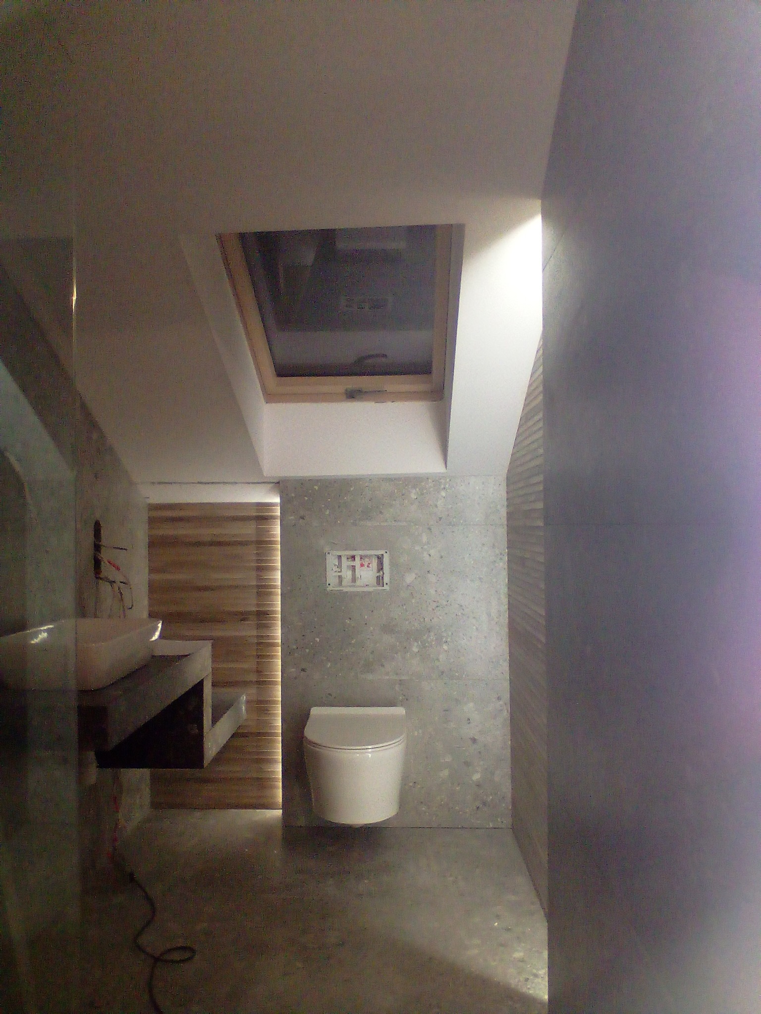 Nowoczesna łazienka z betonowymi ścianami, wiszącą toaletą, umywalką na betonowym blacie i oknem dachowym, widok z góry na niedokończone wykończenie.