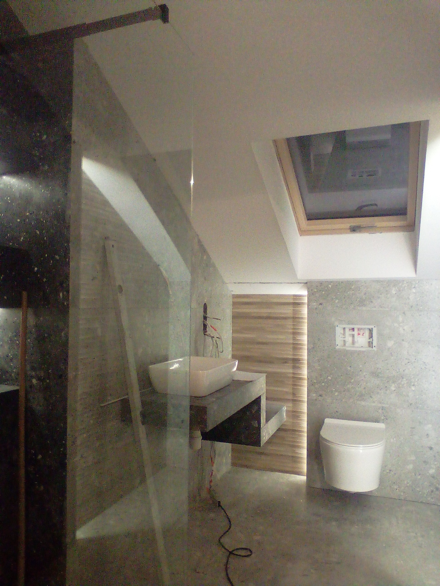 Nowoczesna łazienka z umywalką nablatową na betonowym blacie, wiszącą toaletą, oknem dachowym i szarą mozaiką na ścianach, częściowo przesłonięta szklaną ścianką.