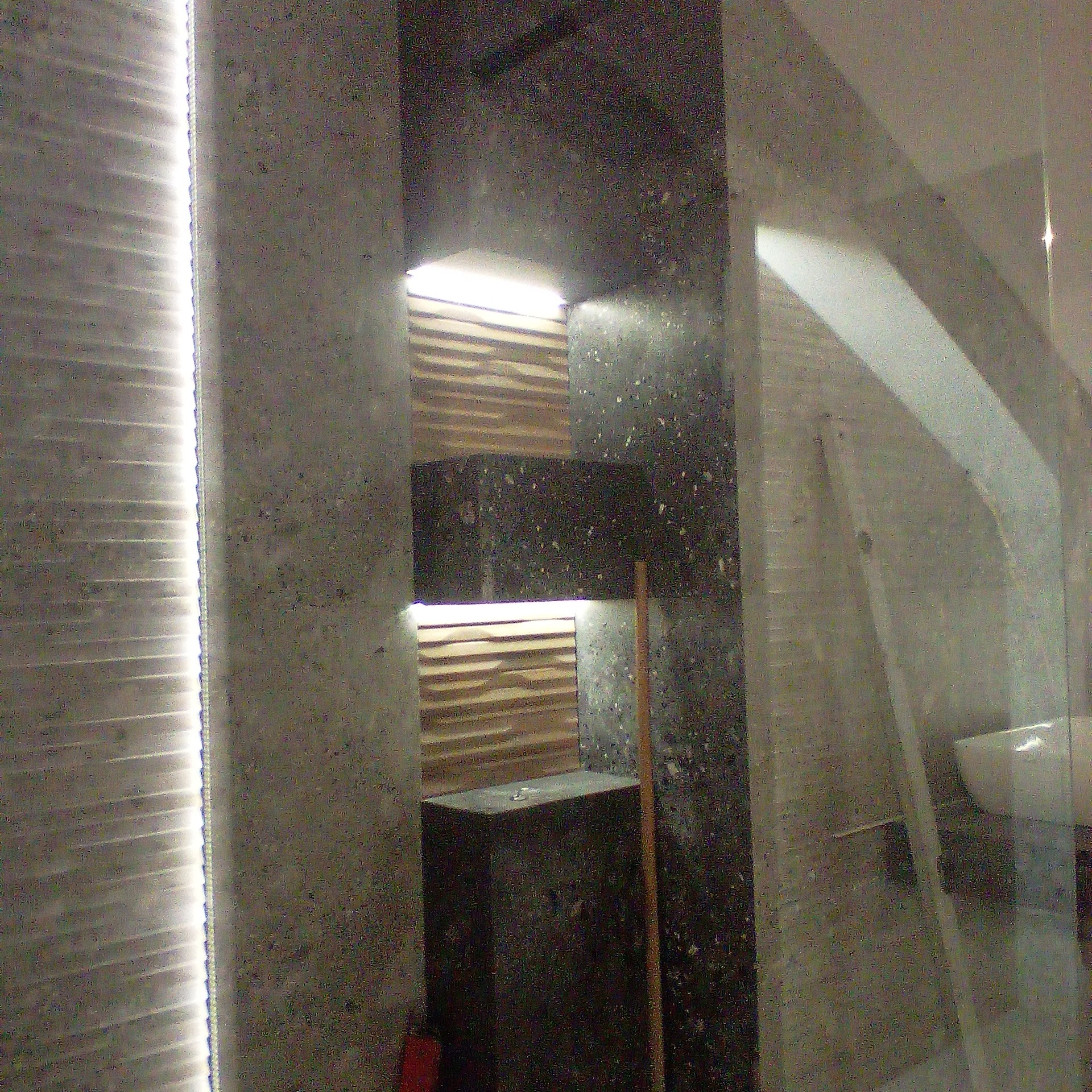 Nowoczesna łazienka z szarą, teksturowaną ścianą, oświetleniem LED wzdłuż krawędzi i drewnianymi panelami dekoracyjnymi, widoczna część umywalki za szklaną ścianką.