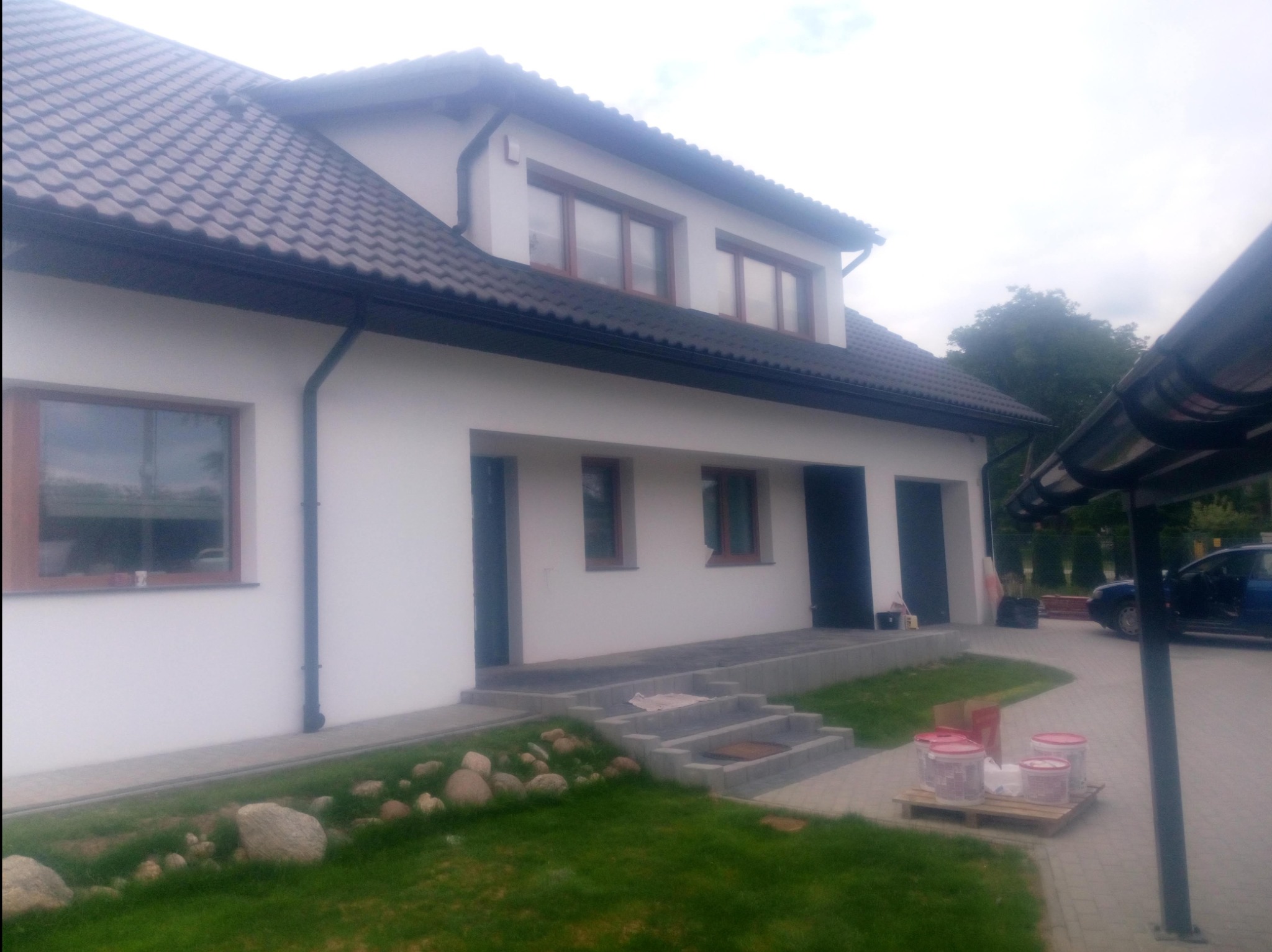 Świeżo ocieplony dom z białymi ścianami, ciemną dachówką i brązowymi ramami okiennymi; widoczne puszki z farbą na palecie.