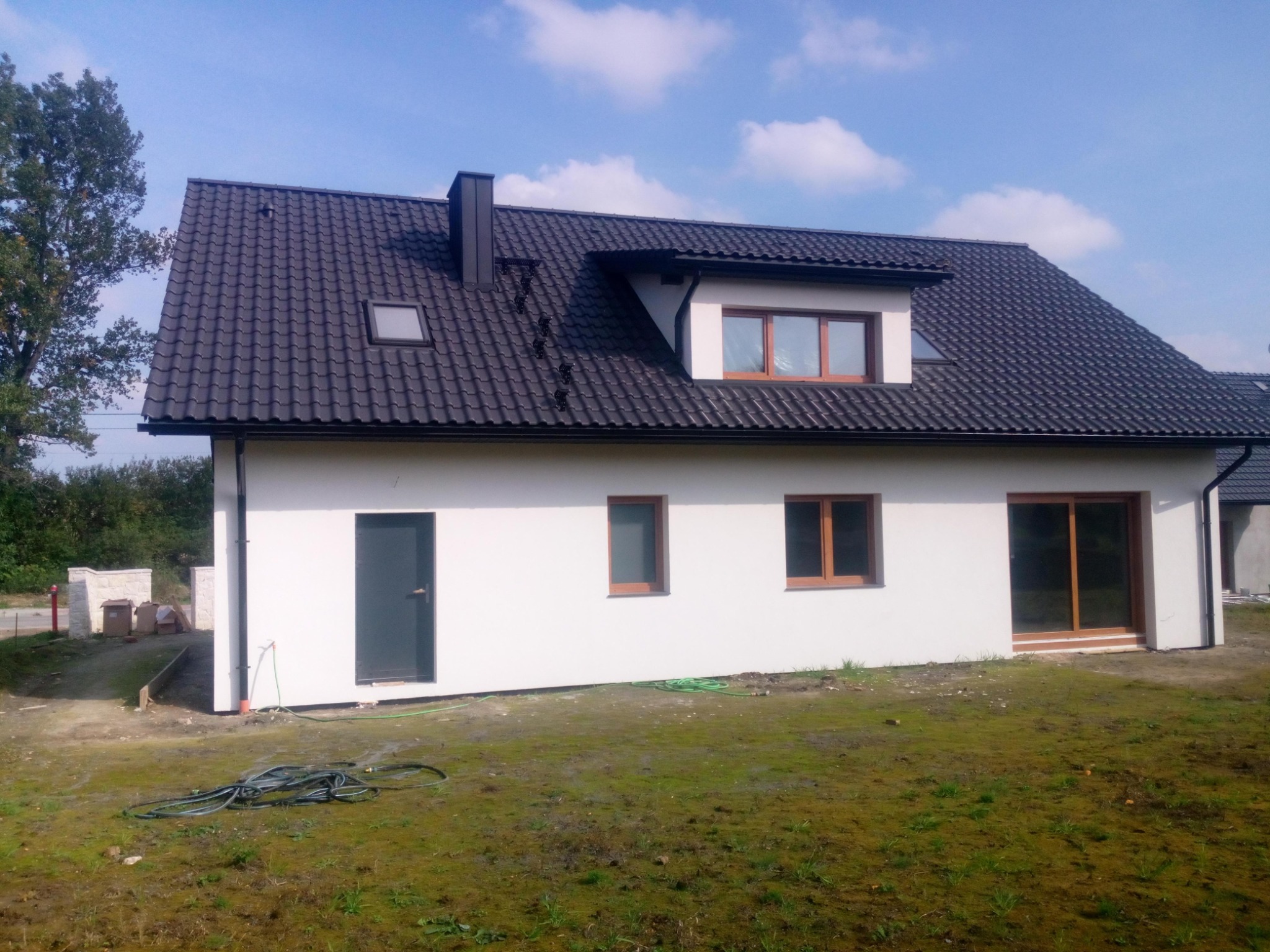 Nowoczesny dom jednorodzinny z ciemną dachówką i białymi ścianami, widok z boku na świeżo zagospodarowaną działkę z zielonym wężem ogrodowym przy ścianie.