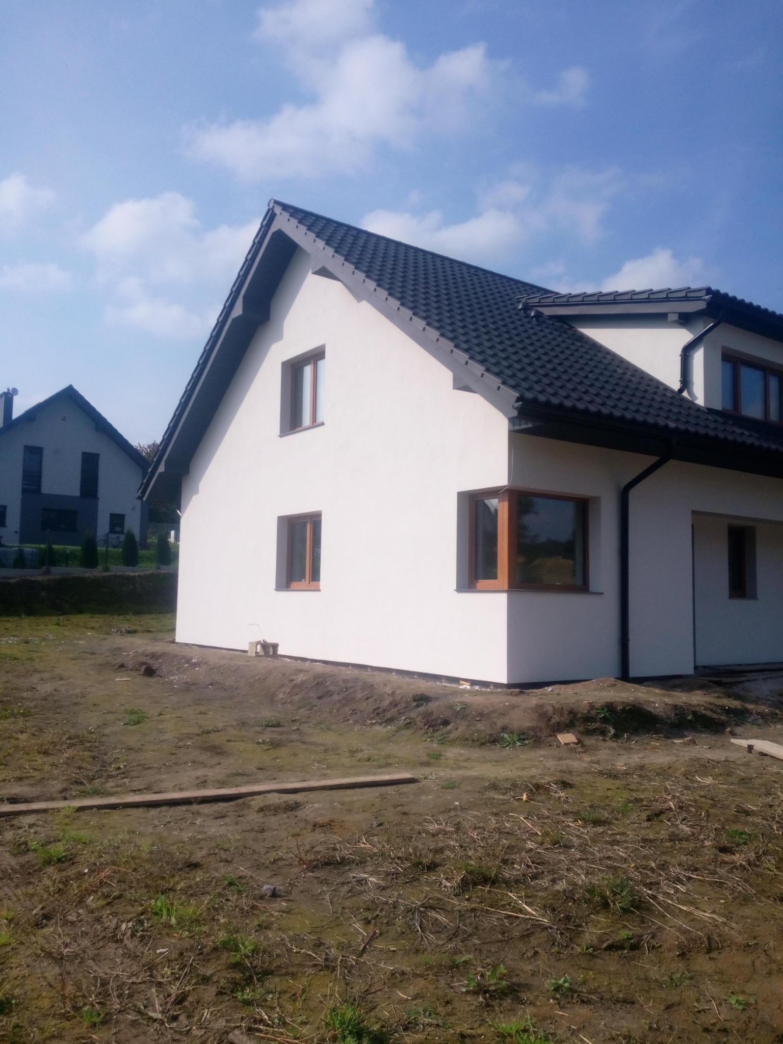 Nowo ocieplony dom jednorodzinny z białymi ścianami i ciemną dachówką, widoczne brązowe ramy okienne, teren wokół domu jeszcze niezagospodarowany.