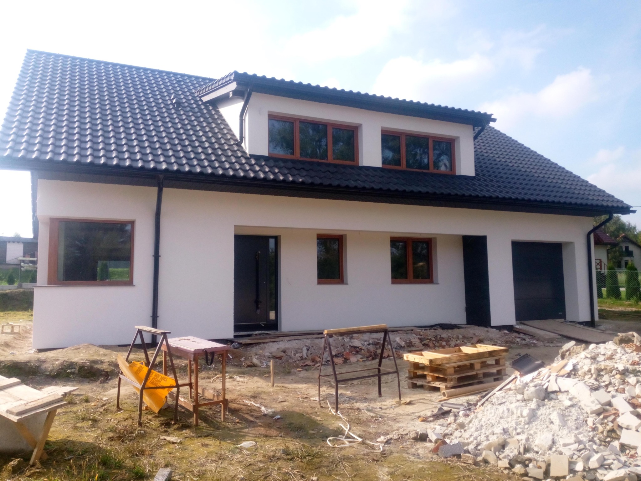 Dwukondygnacyjny dom z ciemną dachówką i białymi ścianami, widoczne rusztowania i gruz sugerujące prace wykończeniowe.