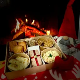 Pudełko z potrawami świątecznymi: pierogi, pasztet z czerwoną wstążką, sałatka jarzynowa, pieczywo i bigos, ustawione na kocu z motywem renifera, w tle kominek i pluszowy skrzat.