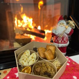 FIRMOWA BUŁA - Świąteczny zestaw dań w tekturowym pudełku: pierogi z kapustą i grzybami posypane skwarkami, krokiety, kapusta z grzybami i suszoną śliwką, dekoracja z pomarańczy i szyszki, w tle płonący kominek...