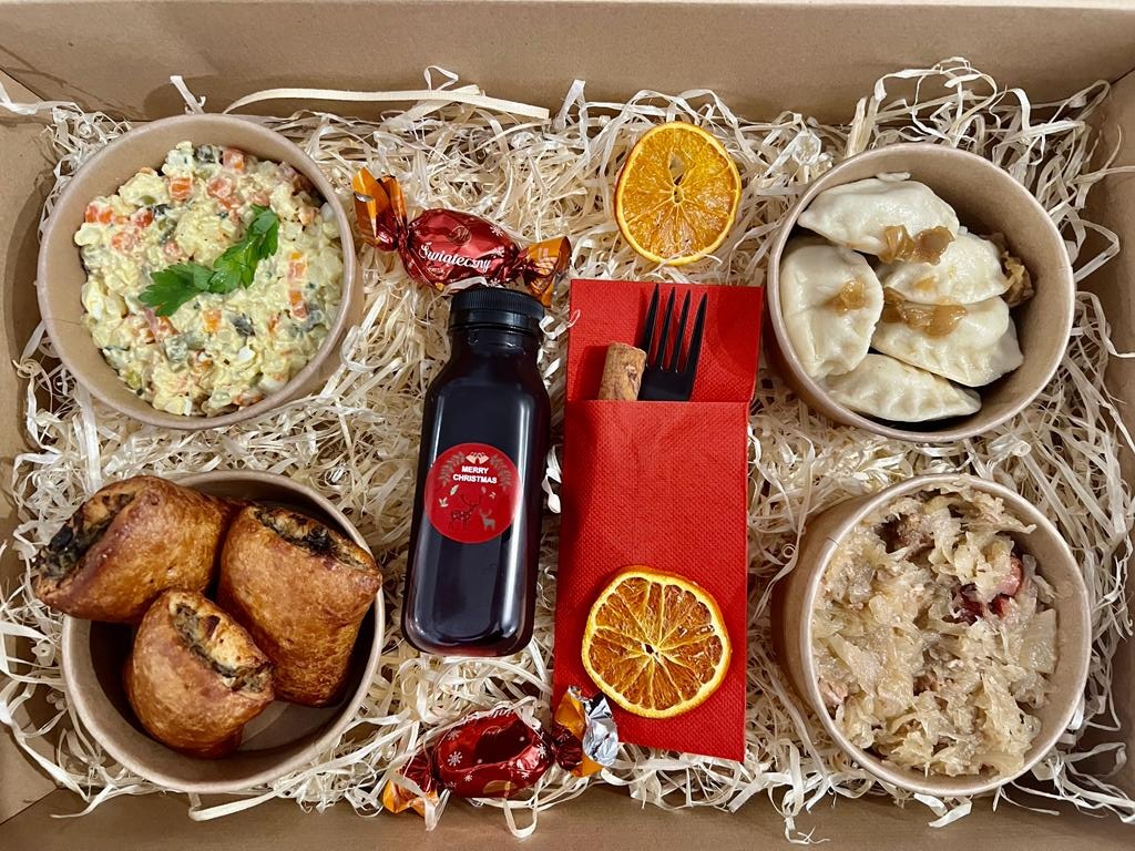 Świąteczne dania w papierowych miseczkach ułożone w kartonowym pudełku na białym sianku: sałatka jarzynowa z natką pietruszki, pierogi z cebulką, kapusta z grzybami, paszteciki, butelka ciemnego...