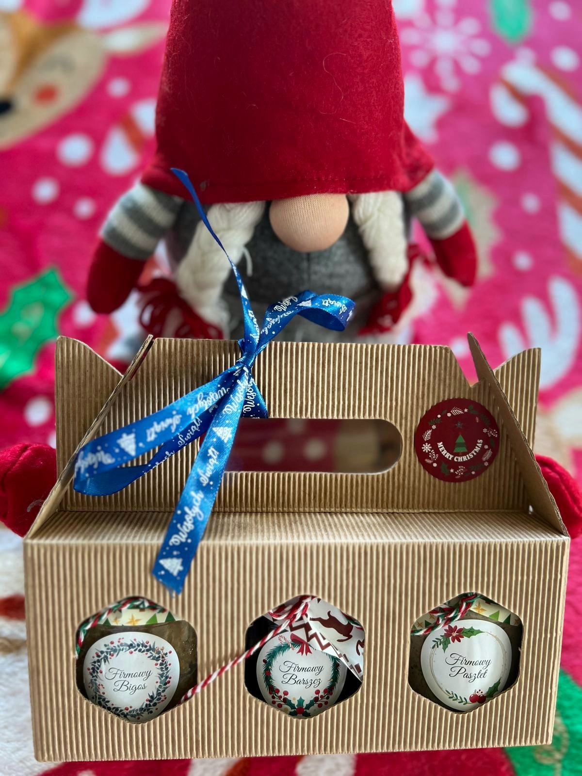 Kartonowe pudełko z trzema słoiczkami firmowych potraw świątecznych (bigos, barszcz, pasztet) z naklejkami i niebieską wstążką z napisem 'Wesołych Świąt', na tle pluszowego krasnala w czerwonej...