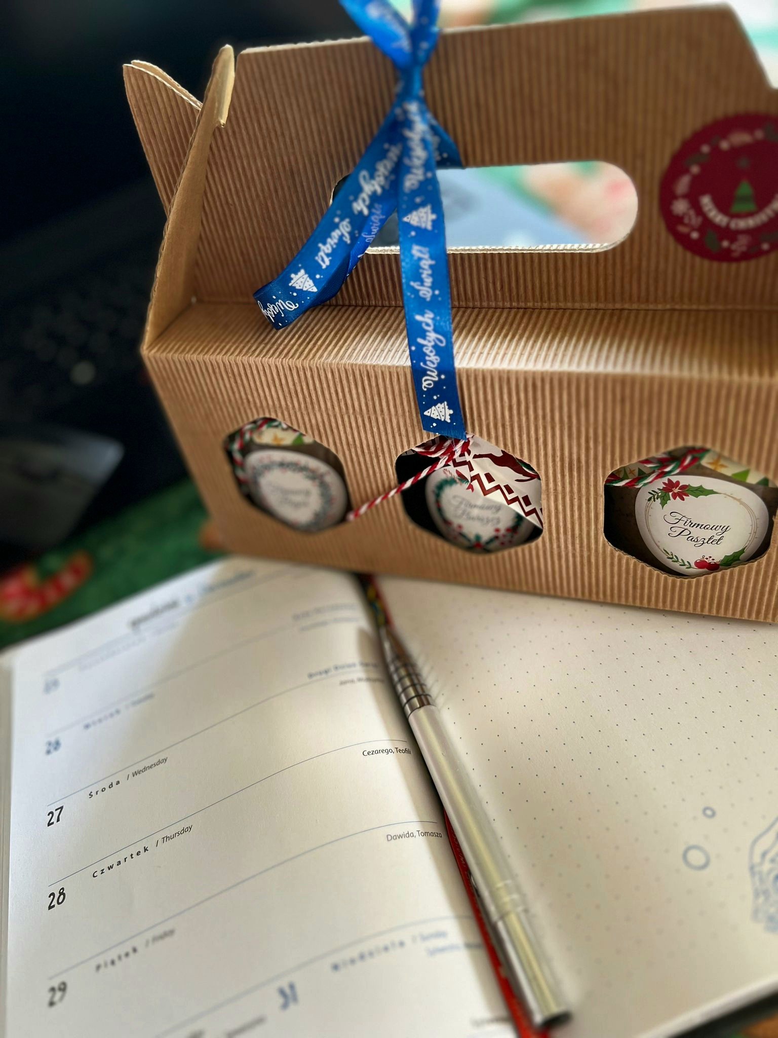 Kartónowe pudełko z trzema okienkami, przez które widać owalne etykiety z napisem 'Firmowy Pasztet', przewiązane niebieską wstążką z napisem 'Wesołych Świąt', leżące na otwartym kalendarzu obok...