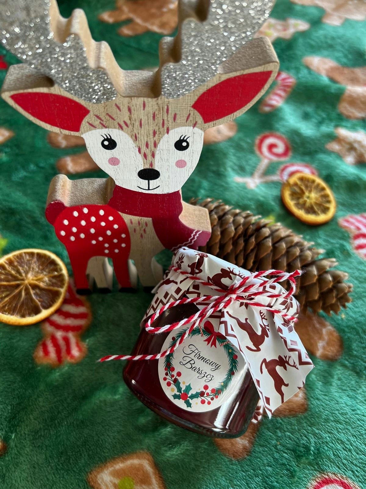 Słoik z firmowym barszczem, ozdobiony świąteczną etykietą i sznurkiem, stoi obok drewnianego renifera z brokatowymi rogami i suszonych plasterków pomarańczy na zielonym kocu w świąteczne wzory.