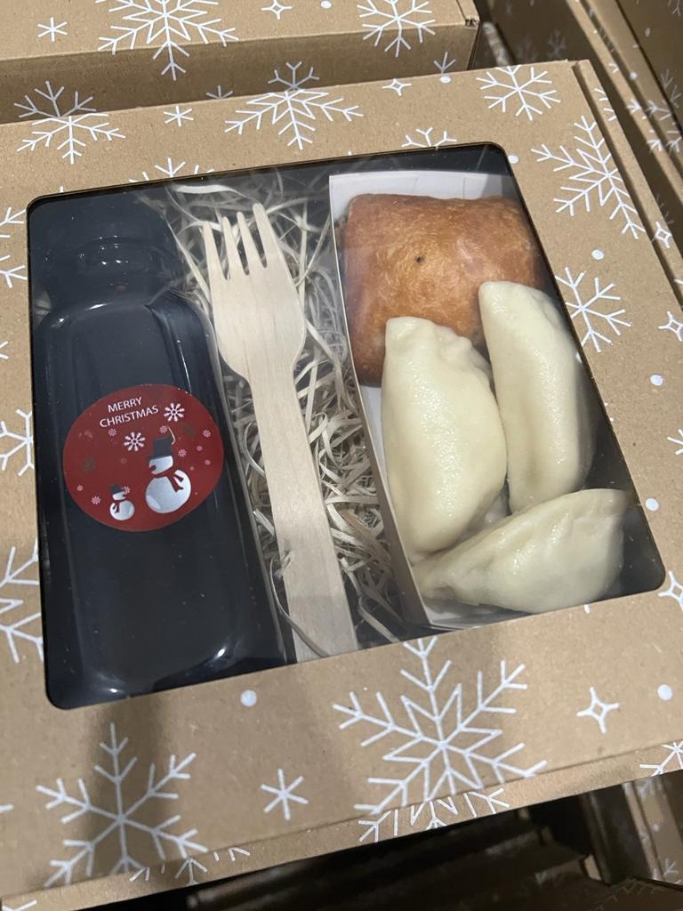 Pudełko z pierogami, krokietem, sosem w butelce z etykietą 'Merry Christmas' i drewnianym widelcem, zapakowane w karton z motywem śnieżynek.