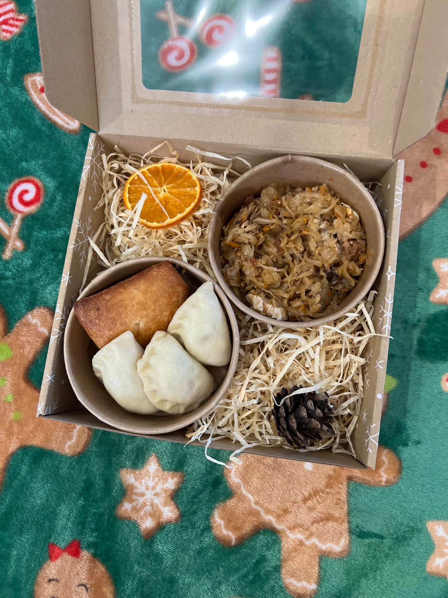 Pudełko z okienkiem wypełnione świątecznymi potrawami: pierogi, kapusta z grzybami, suszona pomarańcza i szyszka, ułożone na zielonym kocu z motywem pierników.