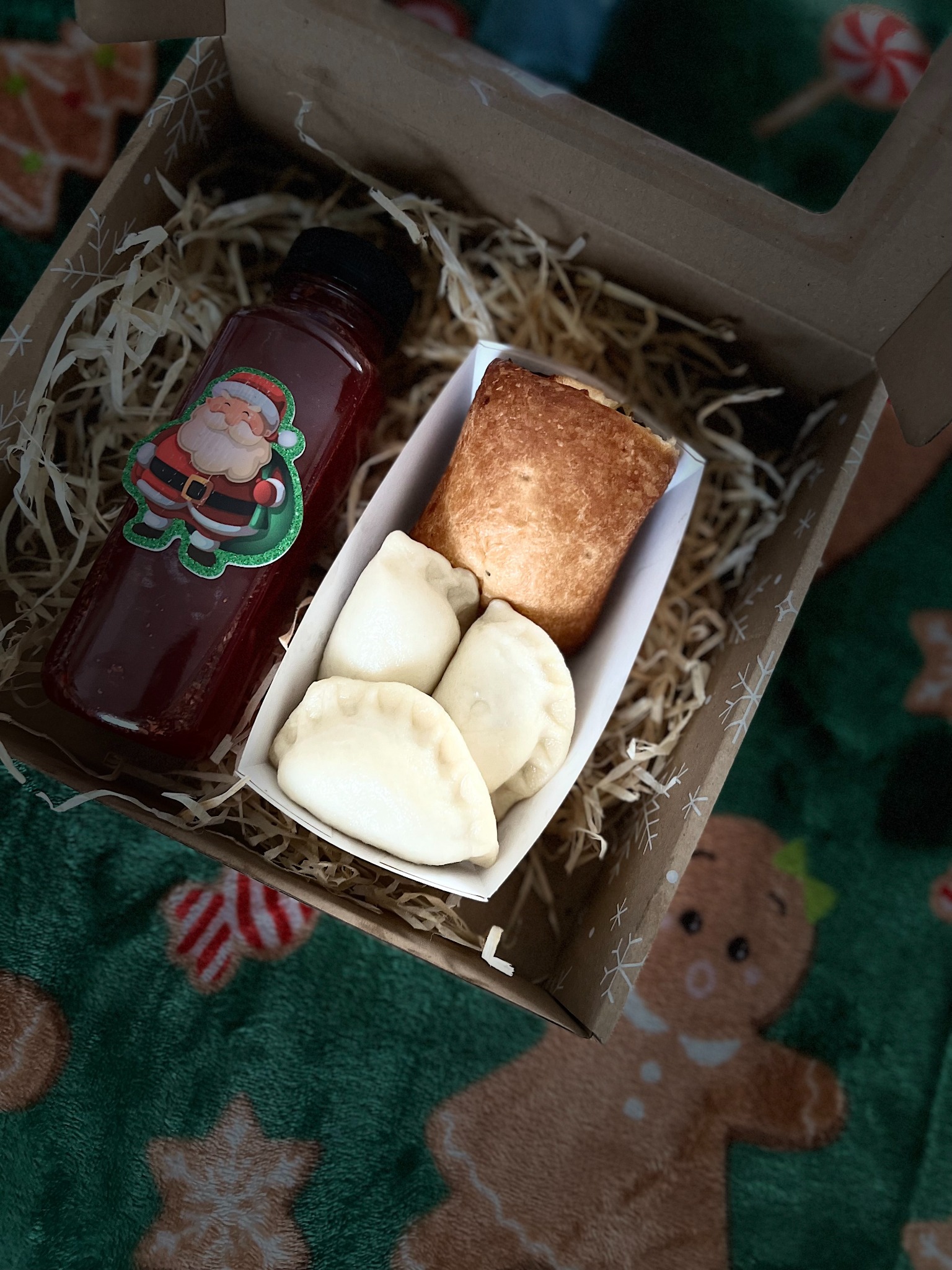 Pudełko z wigilijnym zestawem na wynos: trzy pierogi, pasztecik w papierowej foremce i butelka czerwonego napoju ze świąteczną naklejką Świętego Mikołaja, całość na zielonym tle z motywem pierników.