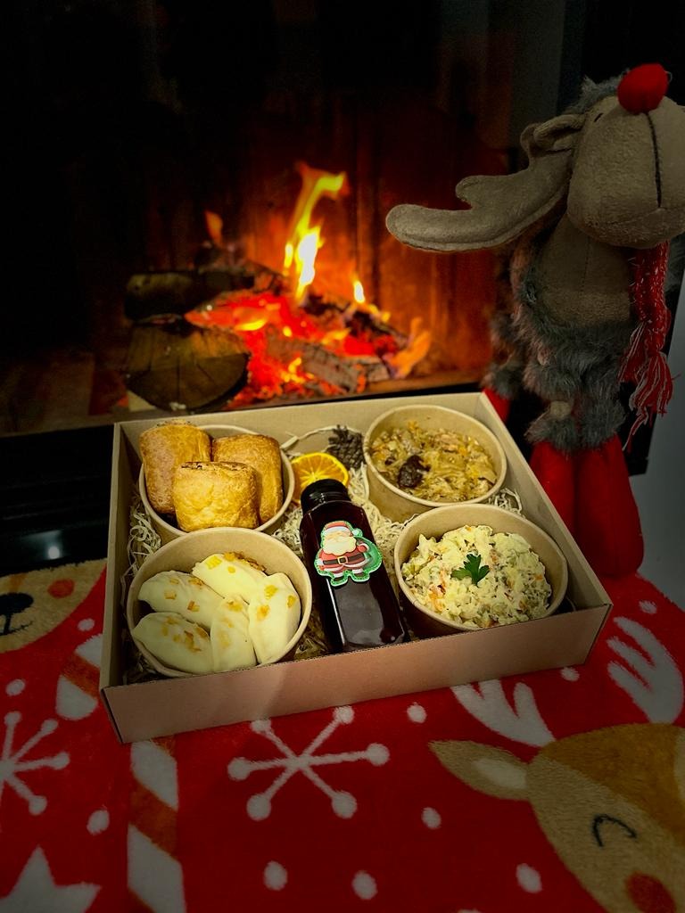Świąteczny catering w Zielonej Górze: pudełko z pierogami, sałatką jarzynową, pasztetem i napojem z etykietą Świętego Mikołaja, ustawione na czerwonym kocu w renifery, z kominkiem i pluszowym...