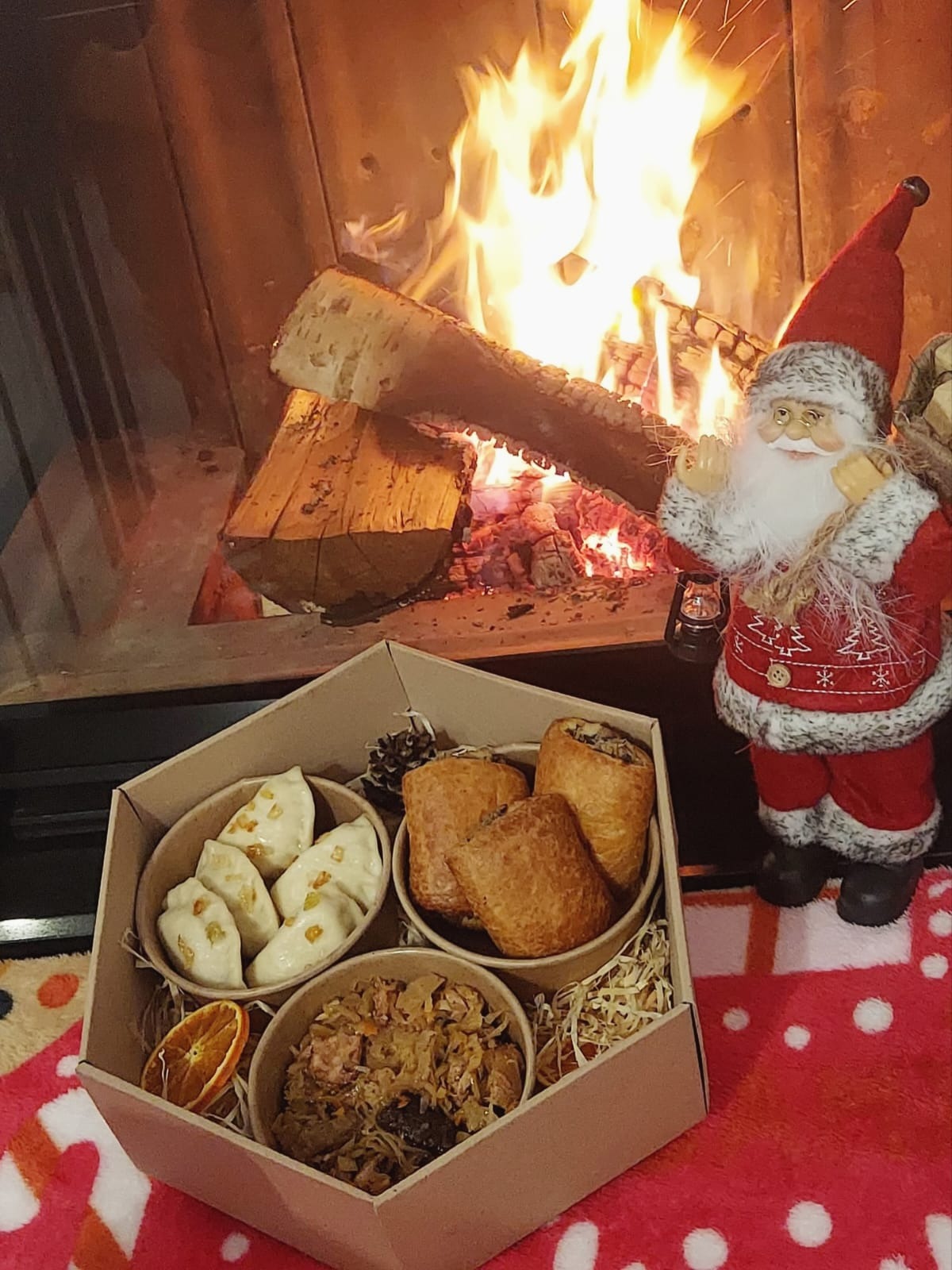 Świąteczny catering w kartonowym pudełku z pierogami, pasztecikami i kapustą z grzybami, na tle płonącego kominka i figurki Świętego Mikołaja z lampionem.