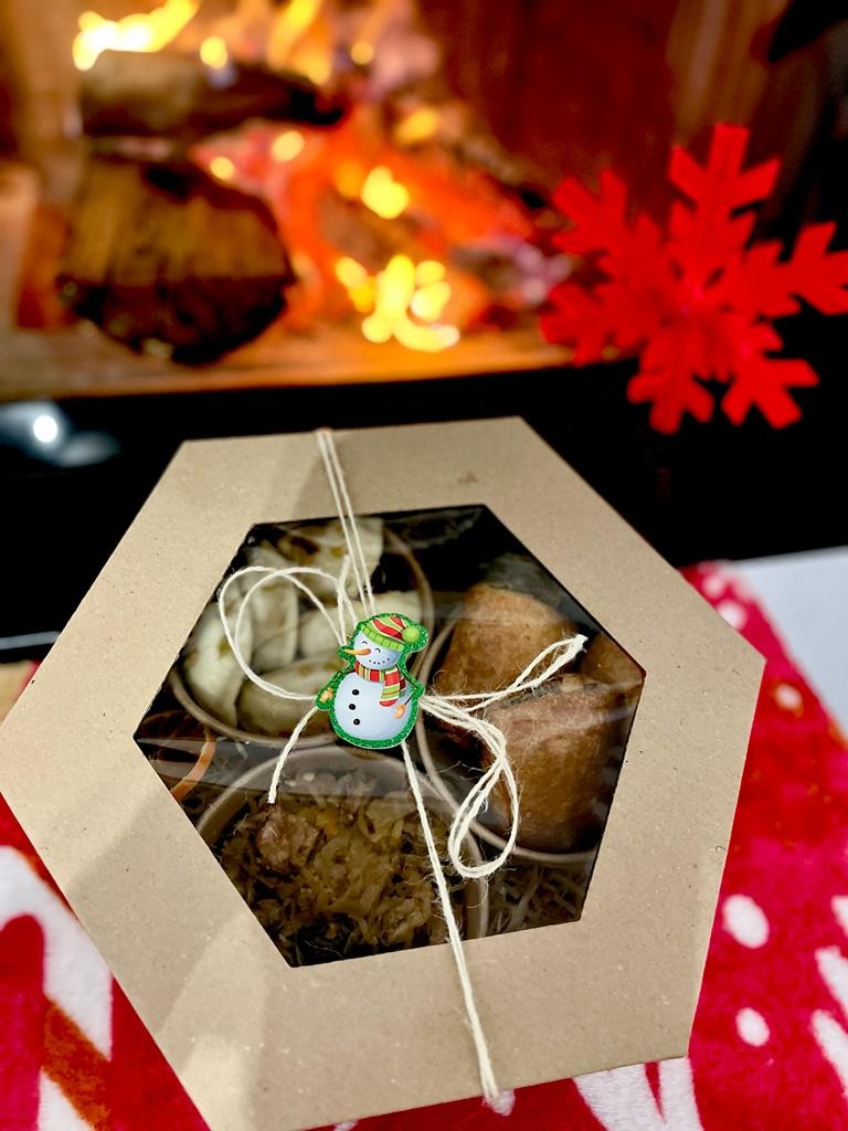 Pudełko z pierogami, kapustą i grzybami oraz pasztecikami, ozdobione sznurkiem i przypinką w kształcie bałwanka, na tle płonącego kominka i czerwonej dekoracji w kształcie śnieżynki.