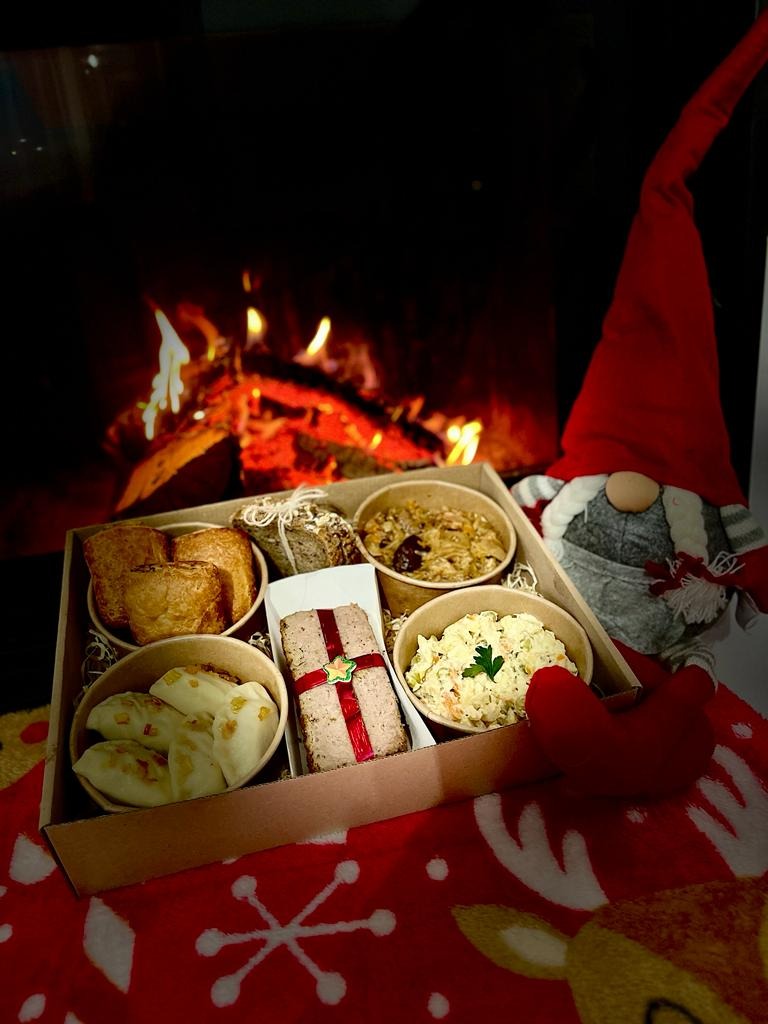 Pudełko z potrawami świątecznymi: pierogi, pasztet z czerwoną wstążką, sałatka jarzynowa, pieczywo i bigos, ustawione na kocu z motywem renifera, w tle kominek i pluszowy skrzat.