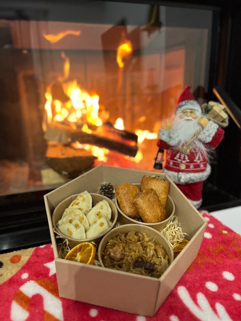 Świąteczny zestaw dań w tekturowym pudełku: pierogi z kapustą i grzybami posypane skwarkami, krokiety, kapusta z grzybami i suszoną śliwką, dekoracja z pomarańczy i szyszki, w tle płonący kominek...