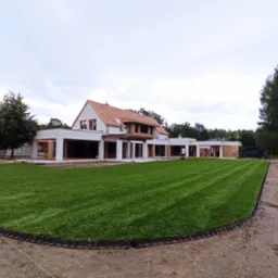 Rozległy trawnik z rolki przed nowoczesnym domem w trakcie budowy, widoczne obrzeża trawnika i przygotowany teren pod dalsze prace.