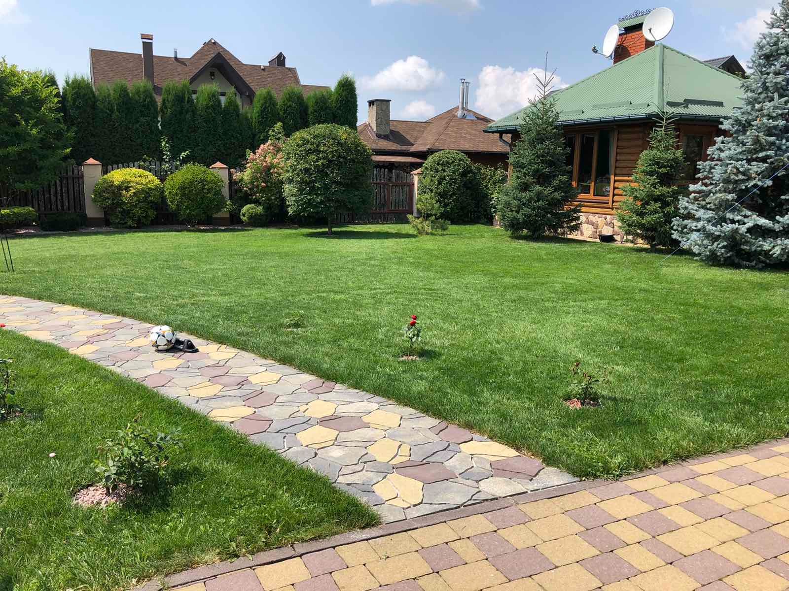 Utrzymany trawnik z kostką brukową w mozaikę, piłka i sandały leżące na ścieżce, zadbane krzewy i drzewa iglaste wokół domu.