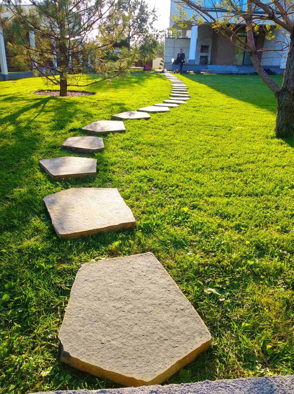 Ścieżka z nieregularnych kamiennych płyt prowadząca przez trawnik w słonecznym ogrodzie w Warszawie, osoba siedząca na tarasie w tle.