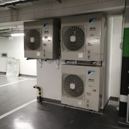 Dan-kan Instalacje Sanitarne Daniel Szewczuk - Trzy jednostki zewnętrzne klimatyzacji Daikin SkyAir zamontowane na ścianie w garażu podziemnym, widoczne rury wentylacyjne pod sufitem, element zabezpieczający ścianę z żółto-czarnymi pasami.