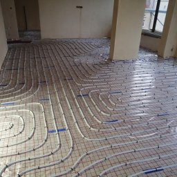 Dan-kan Instalacje Sanitarne Daniel Szewczuk - Białe rury ogrzewania podłogowego ułożone na izolacji z czerwoną siatką, przymocowane niebieską taśmą i czarnymi klipsami, widok z góry w trakcie montażu w surowym wnętrzu.