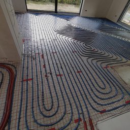 Dan-kan Instalacje Sanitarne Daniel Szewczuk - Rozłożona instalacja ogrzewania podłogowego z niebieskich rurek na folii z czerwoną siatką, widok z góry na surowy stan w nowym domu z dużymi oknami.