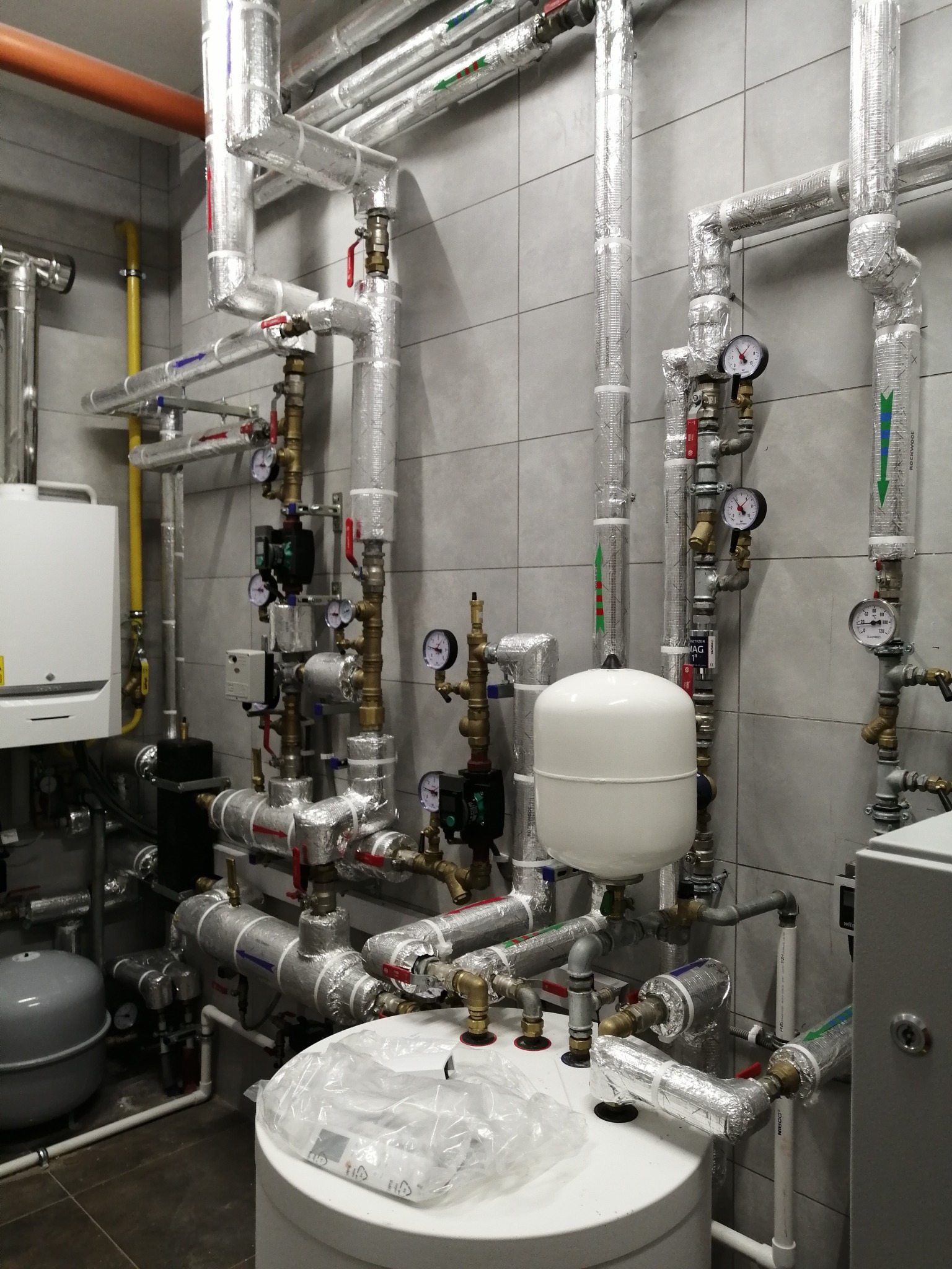 Skomplikowana instalacja rur, zaworów i manometrów w kotłowni, z izolacją termiczną na niektórych odcinkach i białym zbiornikiem wyrównawczym na pierwszym planie, widoczny piec gazowy w tle po...