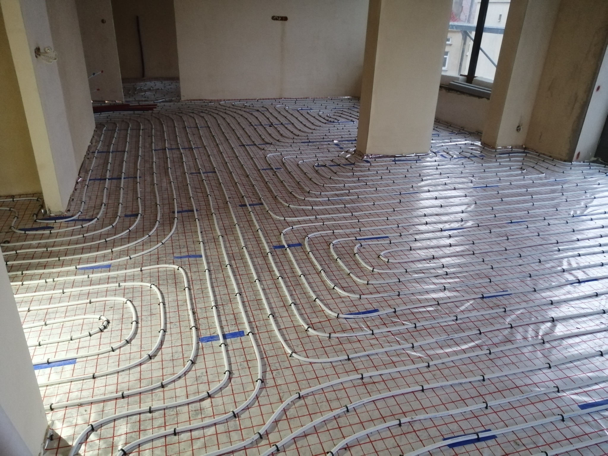 Białe rury ogrzewania podłogowego ułożone na izolacji z czerwoną siatką, przymocowane niebieską taśmą i czarnymi klipsami, widok z góry w trakcie montażu w surowym wnętrzu.