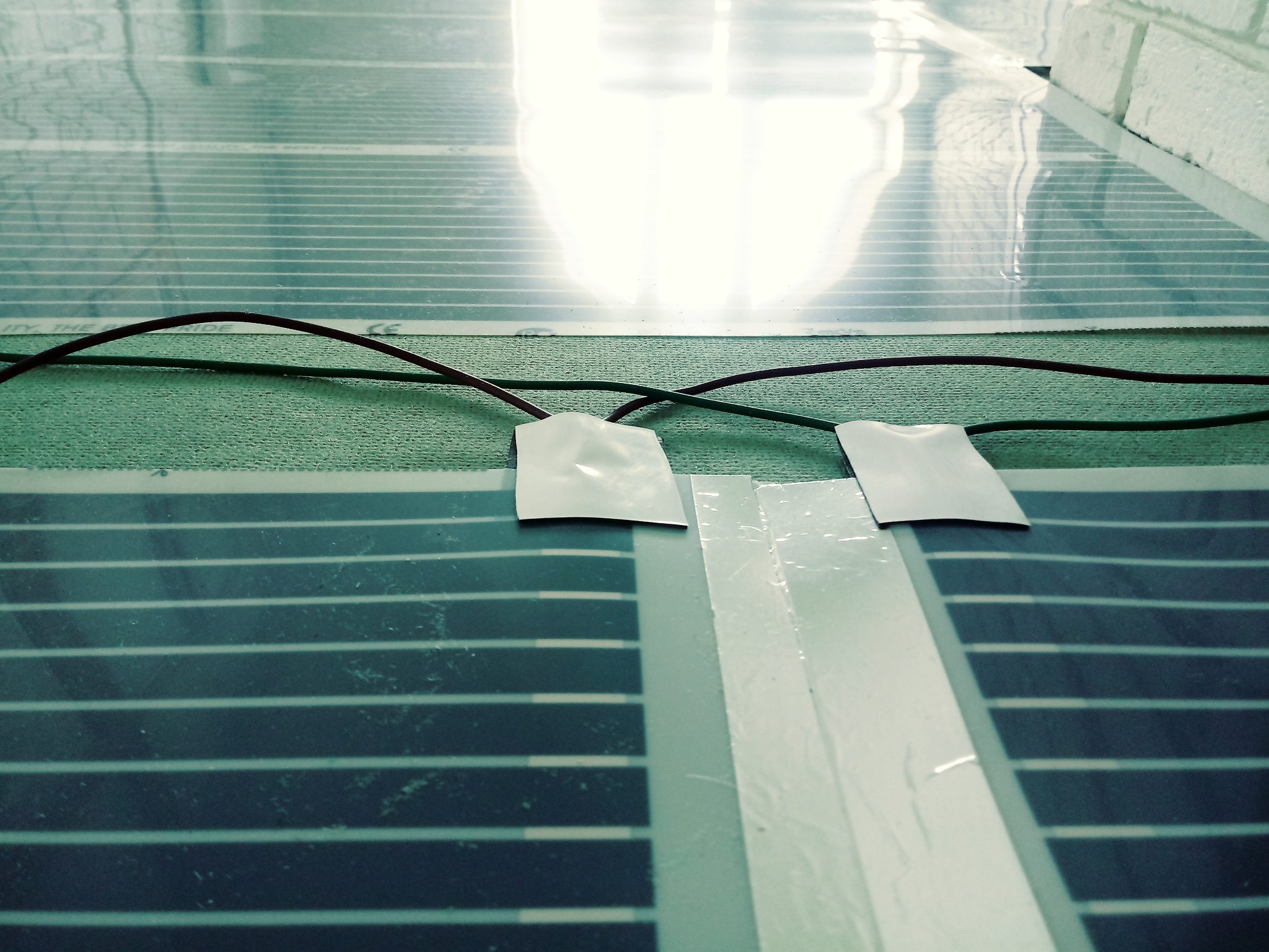 Folia grzewcza na podczerwień rozłożona na podłożu, z widocznymi przewodami i taśmą izolacyjną, przygotowana do montażu ogrzewania podłogowego.