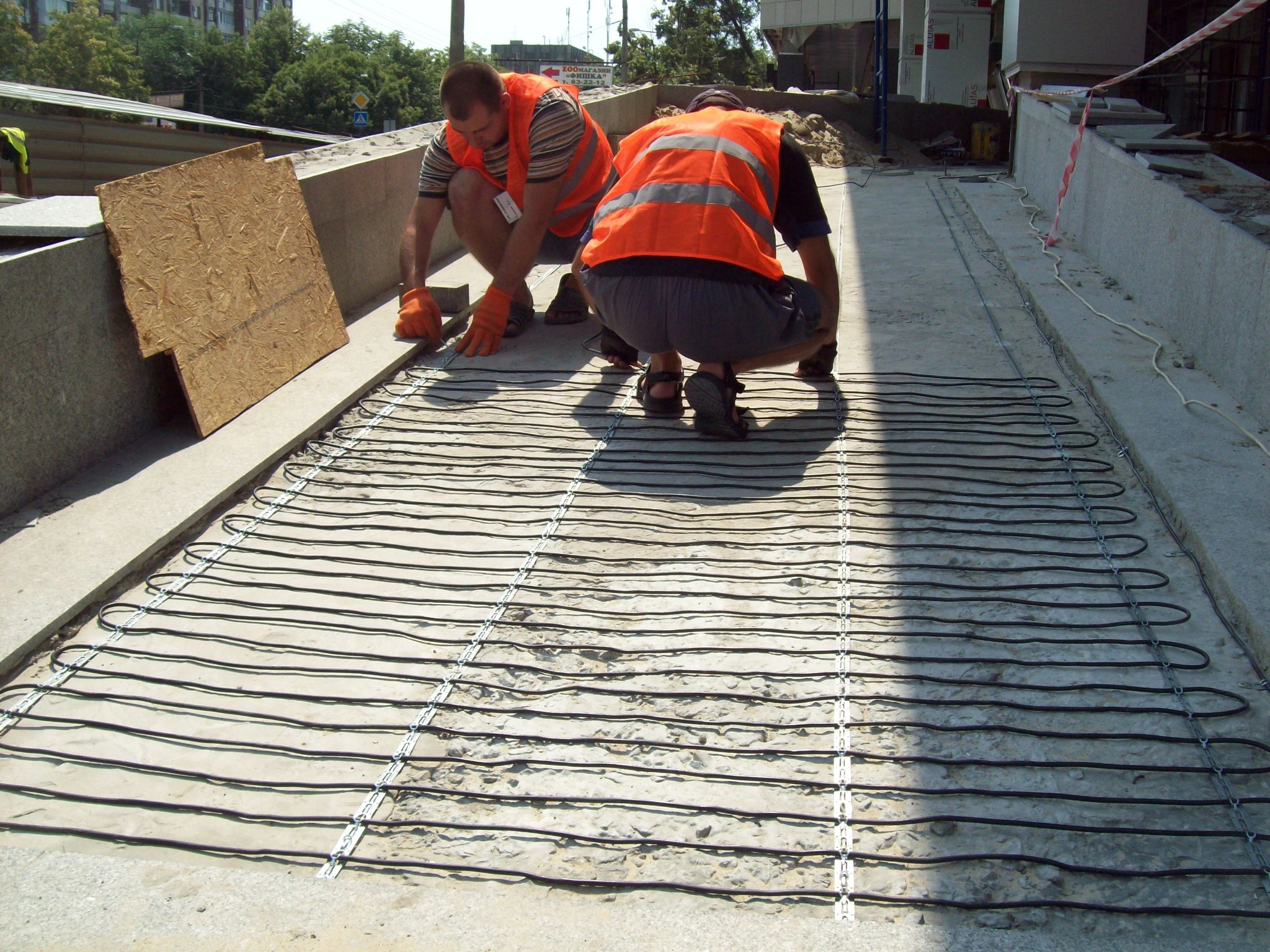 Dwóch pracowników w pomarańczowych kamizelkach instaluje system ogrzewania kablowego na betonowej powierzchni, prawdopodobnie wejście do centrum handlowego; widoczne kable grzewcze przymocowane...