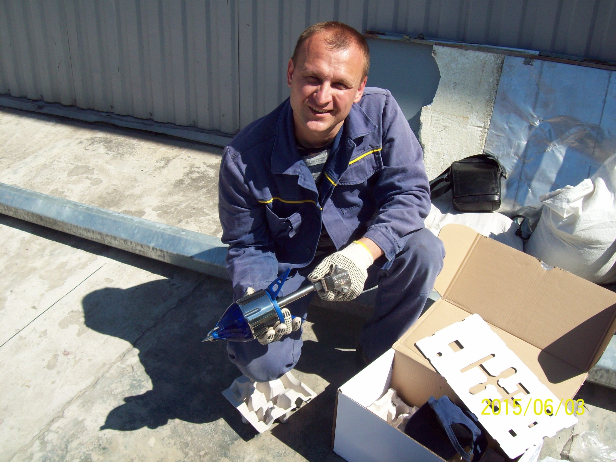 Mężczyzna w roboczym kombinezonie klęczy na dachu, trzyma w rękach narzędzie do instalacji odgromowej, obok kartonowe pudełka i torba.