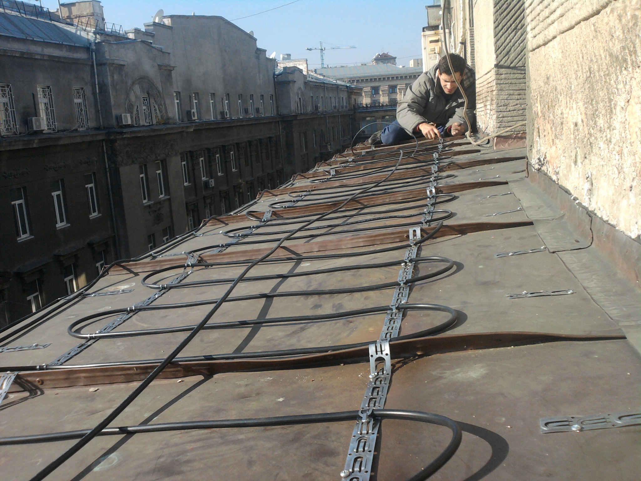Pracownik w kurtce montuje przewód grzejny na dachu budynku z widocznymi starymi kamienicami w tle, używając metalowych uchwytów do mocowania kabla.